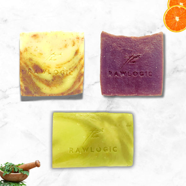Three different colored soap bars from RAWLOGIC on a light background with a sliced orange and mortar and pestle.