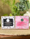 Two soap packages, one labeled 'Charcoal Magic' and the other 'Garden Rose', on a marble surface with decorative elements.