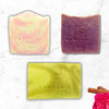 Three bars of Rawlogic soap in different colors on a marble surface with a pink flower and cinnamon sticks.
